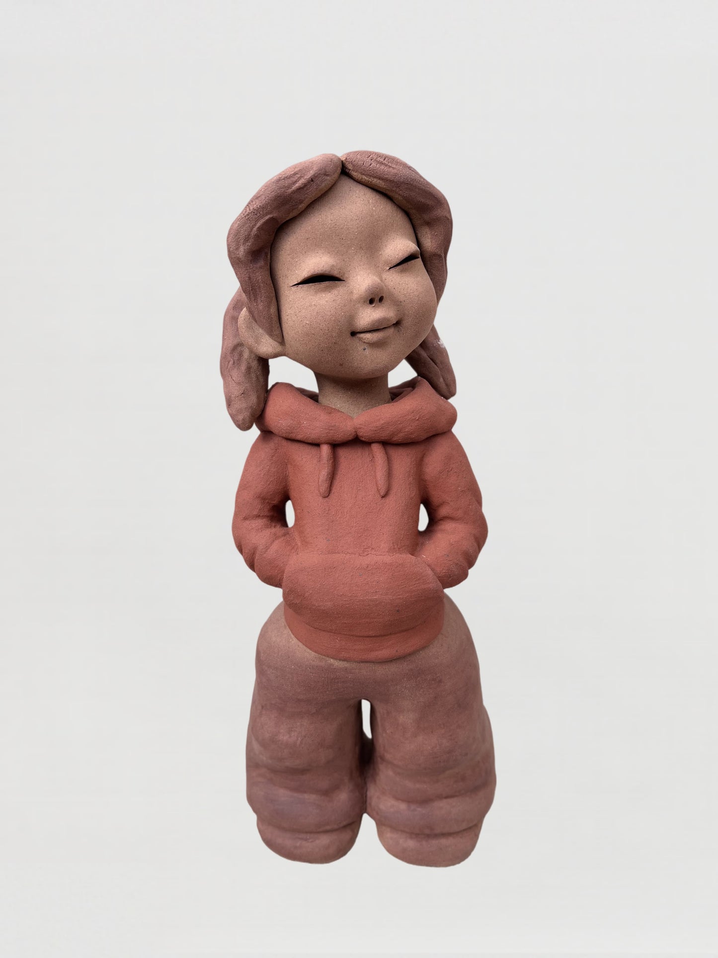 扎著雙辮、雙手插兜站立的微笑女孩陶泥人物雕塑，正面視角，神情安靜自然。