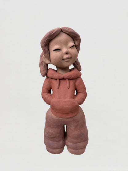 扎著雙辮、雙手插兜站立的微笑女孩陶泥人物雕塑，正面視角，神情安靜自然。